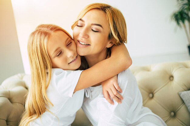 Fröhliche, schöne Mutter und Tochter, die Zeit miteinander verbringen, sich umarmen und Spaß haben, während sie zu Hause auf der Couch sitzen. Familienkonzept