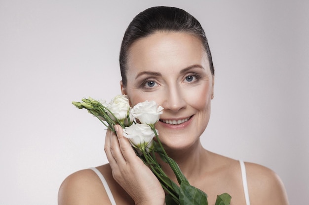 Fröhliche schöne reife Frau mit perfekter Haut und gealterten Falten sind an Blumen mit einem zahnigen Lächeln angelehnt. Hautpflegekonzept, Alterungskosmetik, isoliert auf grauem Hintergrund, Kopierraum, Studioaufnahme, Innenbereich