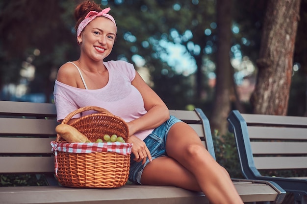 Fröhliche, schöne Rothaarige, die legere Kleidung trägt und mit einem Picknickkorb auf einer Bank in einem Park sitzt.