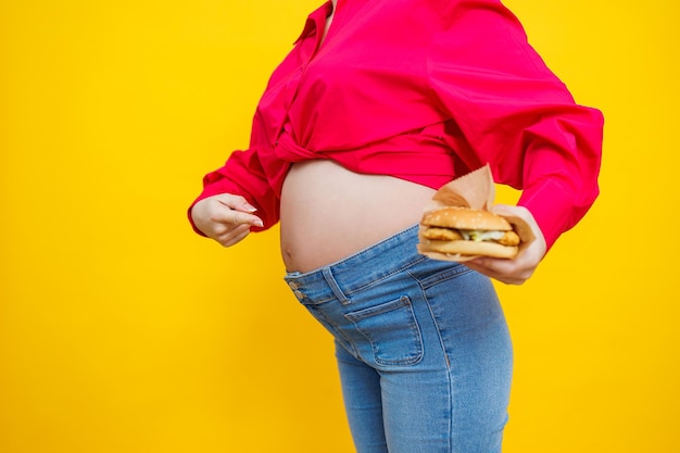 Fröhliche schwangere Frau in rosa Hemd hält Hamburger vor isoliertem gelbem Hintergrund mit überraschtem und schockiertem Gesichtsausdruck. Schädliches Essen während der Schwangerschaft Eine schwangere Frau isst Fast Food