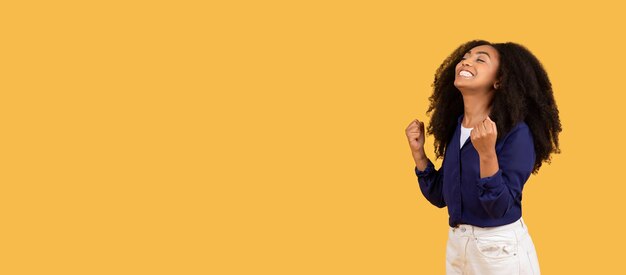 Fröhliche schwarze Dame mit lockigem Haar, die ihre Fäuste zusammenzieht, um Erfolg zu feiern auf gelbem Hintergrund mit