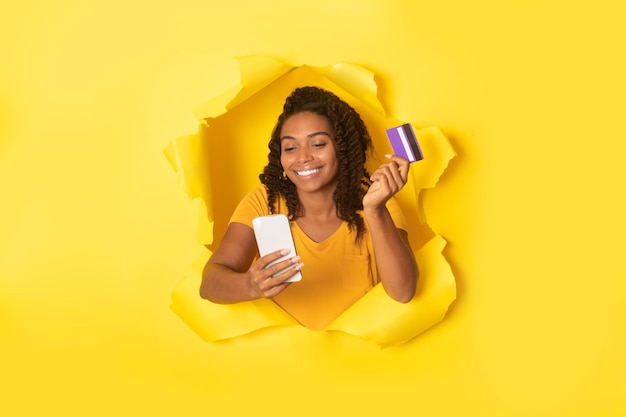 Fröhliche schwarze Frau, die mit Handy und Kreditkarte online bezahlt und in einem zerrissenen Loch aus Papierhintergrund posiert