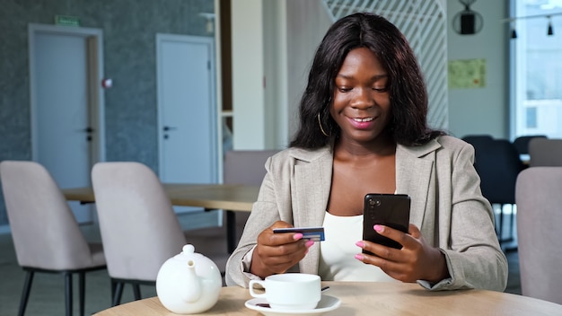 Fröhliche schwarze Frau in stylischer Jacke zahlt online mit Kreditkarte und Handy am Tisch mit Teekanne und Tasse in gemütlicher Cafeteria