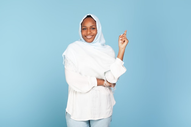 Foto fröhliche schwarze islamische dame im hijab, die mit dem finger nach oben zeigt und einen inspirierenden moment hat, während sie über blauem hintergrund steht
