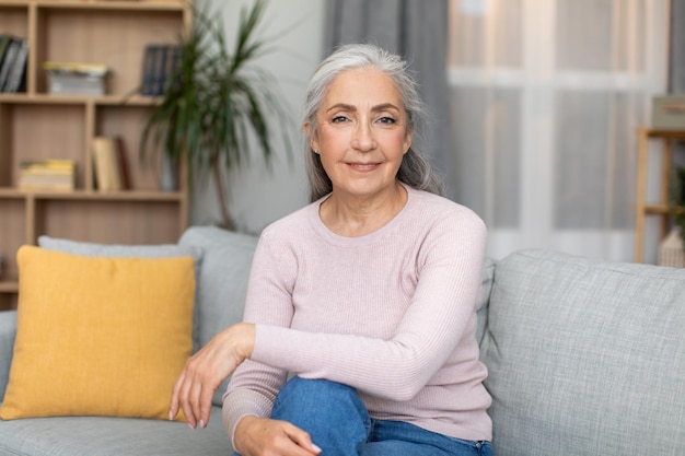 Fröhliche, selbstbewusste, ruhige, hübsche europäische Seniorin mit grauem Haar sitzt auf dem Sofa und genießt Freizeit