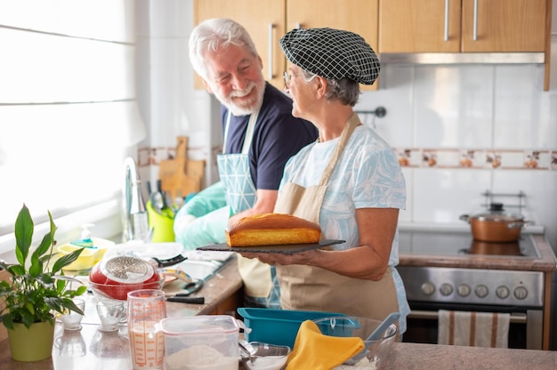 Fröhliche Seniorin, die ihrem Mann beim Abwasch einen frisch gebackenen hausgemachten Pflaumenkuchen zeigt