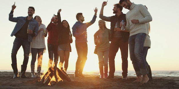 Fröhliche sorglose junge Freunde, die Spaß haben und Bier bei Bonefire am Strand trinken, während die Sonne untergeht