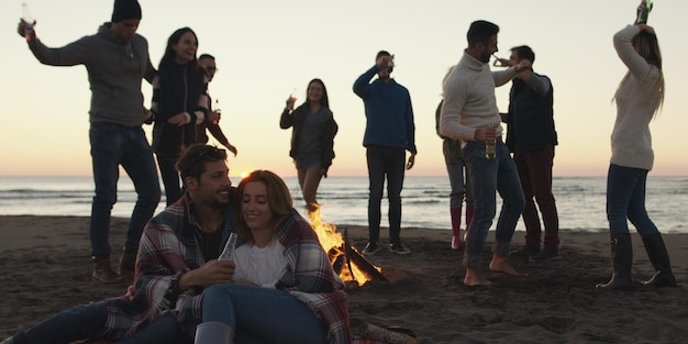 Fröhliche sorglose junge Freunde, die Spaß haben und Bier bei Bonefire am Strand trinken, während die Sonne untergeht