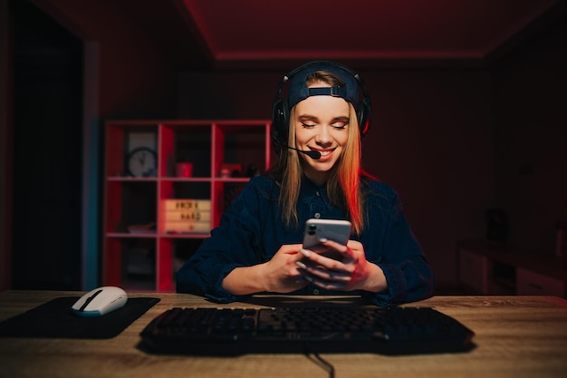 Fröhliche Streamerin in Freizeitkleidung sitzt bei der Arbeit zu Hause am Computer und benutzt ein Smartphone
