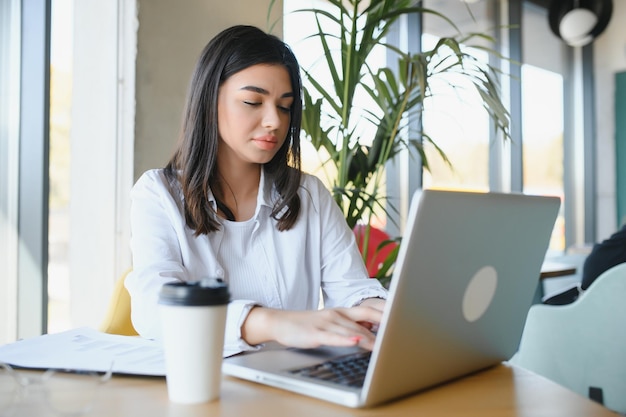 Fröhliche Studentin, die online im Café mit Laptop und Kopfhörern im leeren Raum studiert