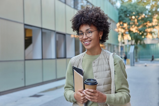 Fröhliche Studentin mit lockigem, buschigem Haar geht auf Straßengetränken Kaffee zum Mitnehmen hält Notizblock und modernes Tablet lächelt freudig