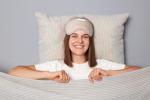 Fröhliche, süße Frau in weißem T-Shirt und schlafender Augenmaske liegt im Bett auf einem Kissen unter einer Decke, isoliert auf grauem Hintergrund und blickt lächelnd in die Kamera, glücklicher Morgen