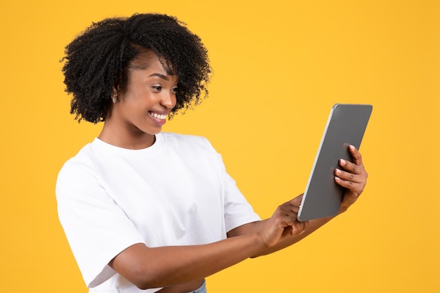 Fröhliche tausendjährige afrikanisch-amerikanische lockige Frau im weißen T-Shirt tippt auf einem Tablet und schaut sich ein Video an