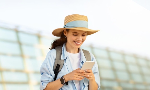Fröhliche tausendjährige kaukasische Touristin mit Hut und Rucksack, die auf der Online-Karte des Telefons tippt