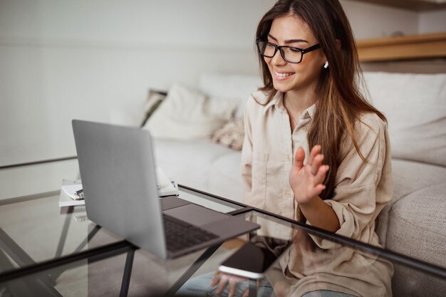 Fröhliche tausendjährige Mischlingsdame mit drahtlosen Kopfhörern und Brille hat Videoanruf auf Laptop mit winkender Hand