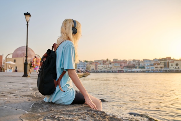 Fröhliche Teenager-Frau in Kopfhörern mit Rucksack, die das Meer der touristischen europäischen Stadt genießt