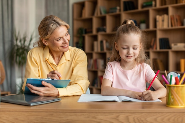 Fröhliche Tutorin, die mit einem intelligenten kleinen Mädchen sitzt, das dem Kind bei den Hausaufgaben hilft und gemeinsam Schulaufgaben vorbereitet