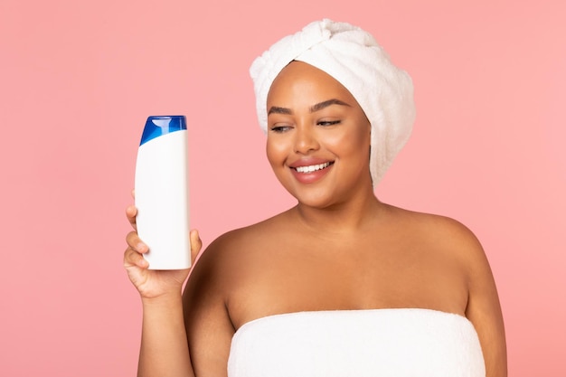 Fröhliche übergewichtige schwarze weibliche Holding-Shampoo-Flasche über rosa Hintergrund