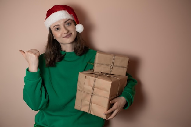 Fröhliche überraschte junge Frau trägt einen Weihnachtspullover Weihnachtsmütze, die zeigt, wie die Hände die Arme zur Seite zeigen, und zeigt auf dem Arbeitsbereich an, der auf einem schlichten pastellfarbenen hellgrünen Hintergrund isoliert ist Frohes neues Jahr 2023 Urlaubskonzept