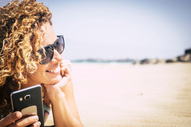 Fröhliche und attraktive junge Frau legte sich auf den weißen Sand am Strand im paradiesischen Ferienort und genieße die Urlaubszeit mit dem Telefon, um mit Freunden zu Hause in Verbindung zu bleiben