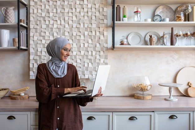 Fröhliche und erfolgreiche Geschäftsfrau im Hijab zu Hause mit Laptop, während muslimische Frau steht