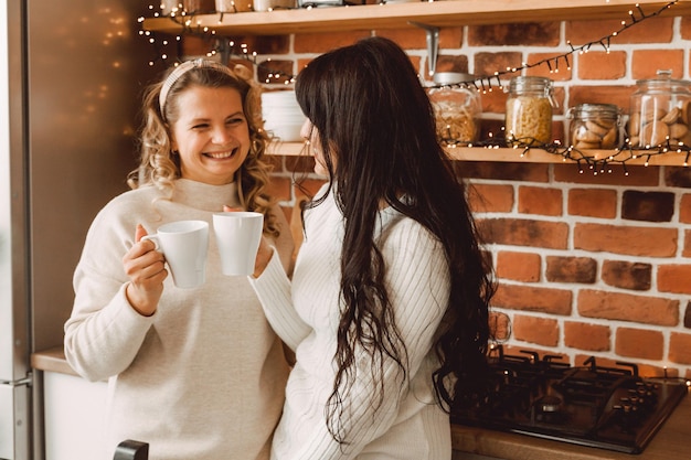 Fröhliche und fröhliche Frauen, die zu Hause in der Küche Kaffee sprechen und trinken. Blondine und Brünette reden miteinander