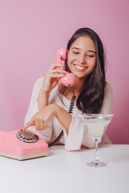 Fröhliche und lächelnde junge brünette Frau mit Getränk, verwendet ein Vintage-Telefon. Porträt auf einer rosa Oberfläche