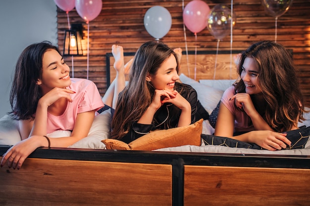 Fröhliche und schöne junge Frauen, die auf dem Bett liegen und sich ansehen. Sie lächeln und lachen. Mädchen tragen Pyjamas. Sie sind in einem Raum.