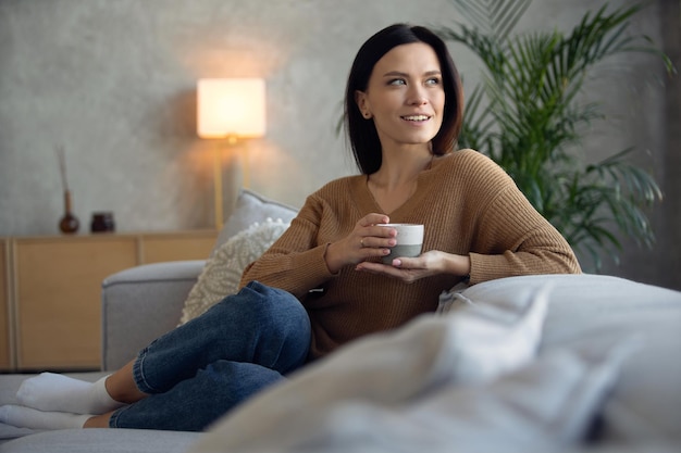 Fröhliche, verträumte Frau mittleren Alters, die auf einem bequemen Sofa im Wohnzimmer mit einer Tasse schwarzem Tee oder Kaffee sitzt