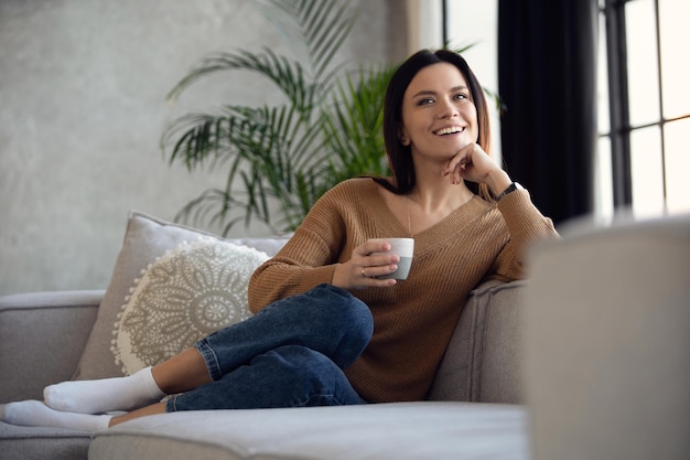 Fröhliche, verträumte Frau mittleren Alters, die auf einem bequemen Sofa im Wohnzimmer mit einer Tasse schwarzem Tee oder Kaffee sitzt