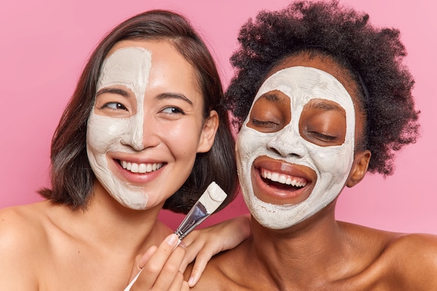 Foto fröhliche, vielfältige frauen tragen gesichtsmasken mit kosmetischem bürstenlächeln auf und zeigen weiße zähne, die sich eng um haut und körper kümmern, die über rosafarbener wand isoliert sind.