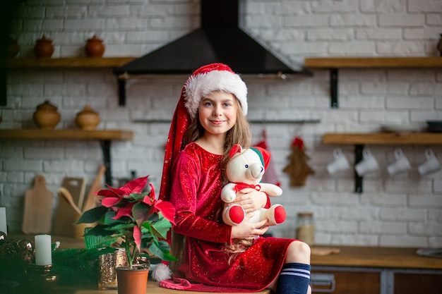 Fröhliche Weihnachten. Nettes kleines Mädchen in Sankt-Hut hält einen Teddybären, der in der Küche sitzt und wartet auf den Feiertag.