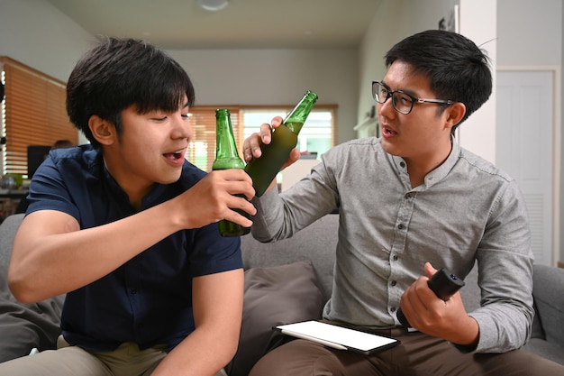 Fröhliche zwei asiatische Männer, die mit ihren Biergläsern klirren, während sie das Fußballspiel auf dem Sofa anfeuern