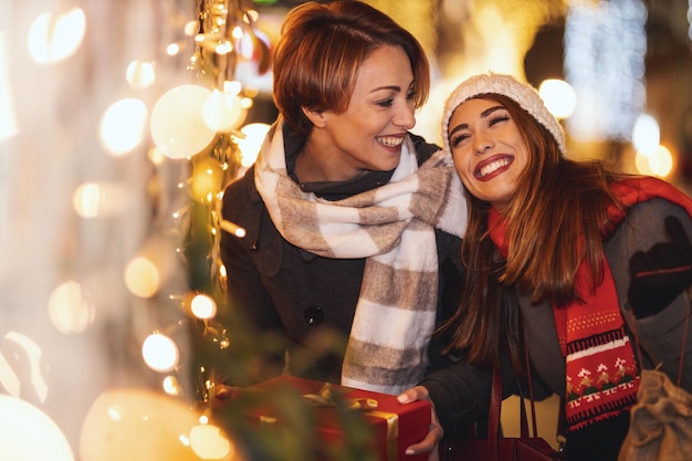 Fröhliche zwei junge Frauen amüsieren sich in der Weihnachtsnacht auf der Stadtstraße. Sie lachen und kaufen Geschenke für ihre Lieblinge.