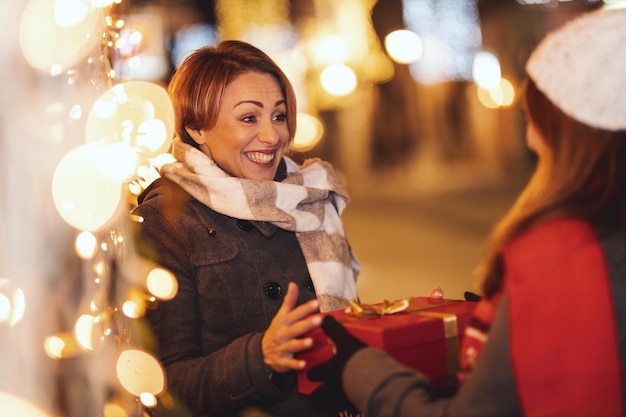 Fröhliche zwei junge Frauen amüsieren sich in der Weihnachtsnacht auf der Straße der Stadt. Sie lachen und beschenken sich gegenseitig.