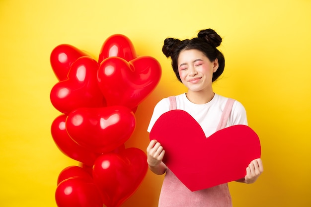 Fröhlichen Valentinstag. Dumme und schöne asiatische Frau, die verträumt lächelt, rotes Herz zeigt, romantisches Datum mit Liebhaber bildend darstellt, auf gelbem Hintergrund stehend.
