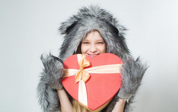 Fröhlichen Valentinstag. Kleine Mädchen tragen Wintermütze Schal. Wintermodetrends für Kinder. Valentinstag. Geschenkbox in Herzform. Liebe. Boxtag. valentinstag einkaufen. mit Liebe präsentieren