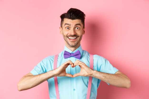 Fröhlichen Valentinstag. Lächelnder lustiger Kerl mit Schnurrbart, der Herzzeichen zeigt und Liebhaber ansieht