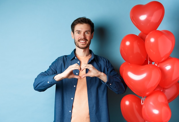 Fröhlichen Valentinstag. Verliebter Freund, der dem Geliebten eine Herzgeste zeigt und lächelt und in der Nähe von roten Luftballons auf blauem Hintergrund steht.
