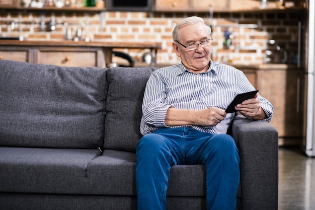 Fröhlicher älterer Mann, der auf der Couch sitzt, während er ein e-Buch liest