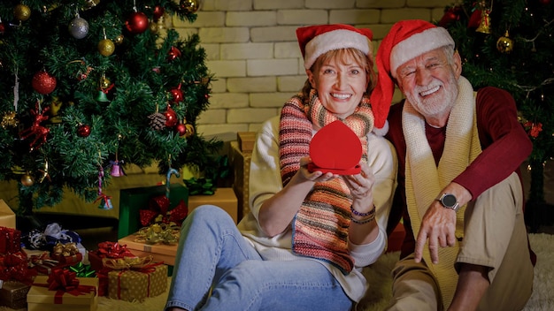 Fröhlicher älterer Mann und Frau in Weihnachtsmützen lächeln und schauen in die Kamera, während sie gemeinsam ein Weihnachtsgeschenk öffnen