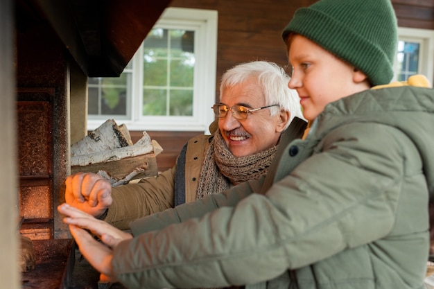 Fröhlicher älterer Mann und sein Enkel stehen am Kamin mit brennendem Brennholz, während sie sich an einem kühlen Herbsttag im Landhaus warm werden