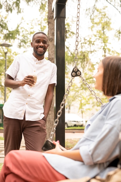 Fröhlicher Afrikaner mit Getränk, der vor einer jungen Frau steht, die sich auf der Schaukel entspannt, während sie am Sommertag im Park mit ihr spricht