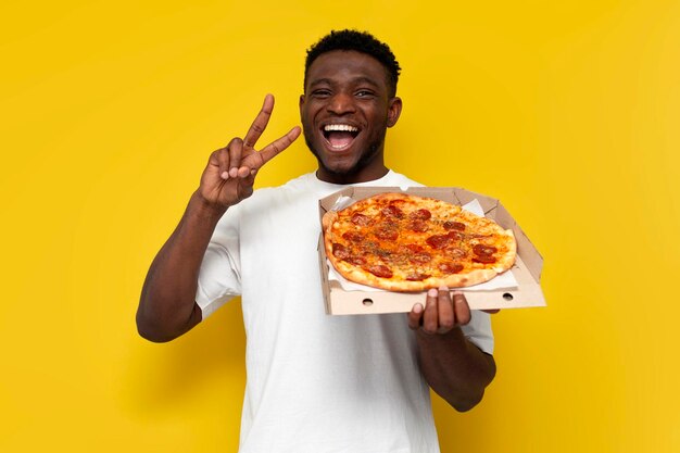 Fröhlicher afrikanisch-amerikanischer Mann in weißem T-Shirt hält eine Pizza-Schachtel und zeigt eine Friedensgest