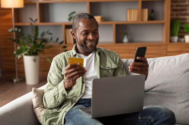 Fröhlicher afrikanisch-amerikanischer Mann mittleren Alters sitzt mit Laptop auf Sofa und nutzt Kreditkarte und Smartphone für Online-Shopping