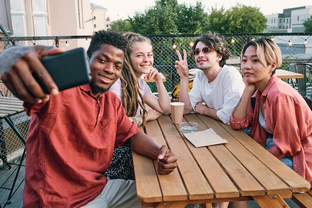 Fröhlicher afrikanischer Kerl macht Selfie mit interkulturellen Freunden am Holztisch