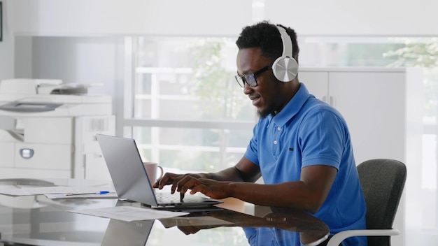 Fröhlicher afrikanischer Manager, der über Kopfhörer Musik hört und am Laptop arbeitet