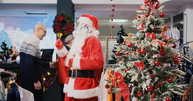 Fröhlicher afroamerikanischer Arbeiter verkleidet als Weihnachtsmann im weihnachtlich verzierten Bekleidungsgeschäft des Einkaufszentrums. Mitarbeiter läuten Weihnachtsglocken und begrüßen glückliche Kunden im festlich geschmückten Modegeschäft