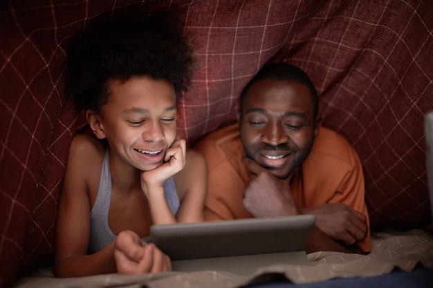 Fröhlicher afroamerikanischer Junge, der mit seinem Vater unter karierter Decke in der Dunkelheit einen aufregenden Film auf einem digitalen Tablet sieht