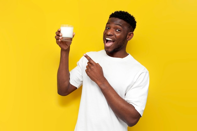 Fröhlicher afroamerikanischer Mann hält ein Glas Milch in der Hand und zeigt darauf auf gelbem, isoliertem Hintergrund